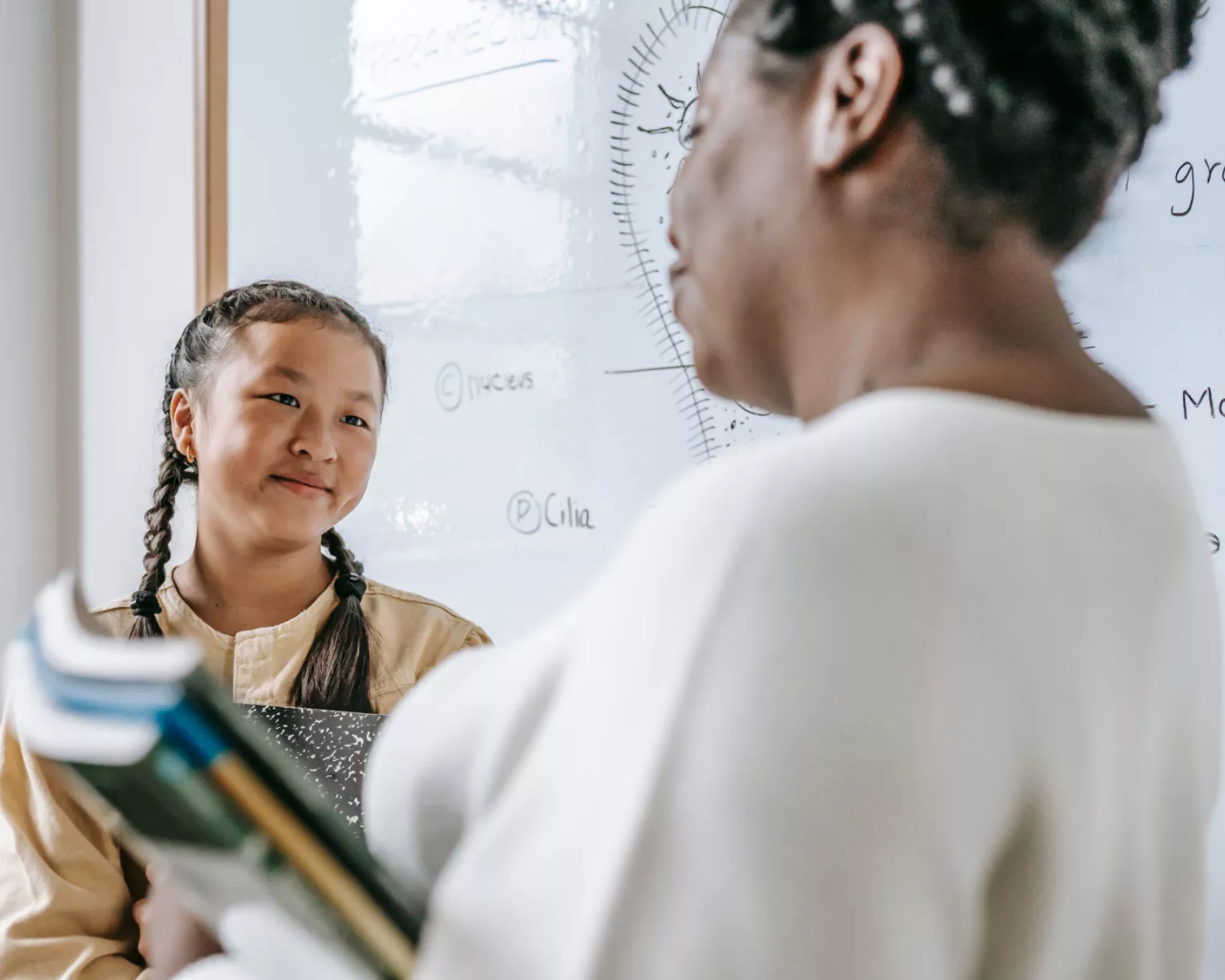 A student with a white dry-erase board covered in math equations behind them smiles at a teacher whose back is to the camera.