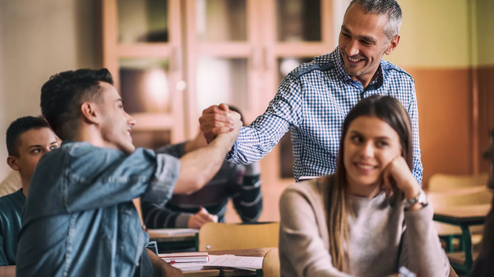 A high school teacher in a plaid shirt smiles as they fist-bump a smiling student.