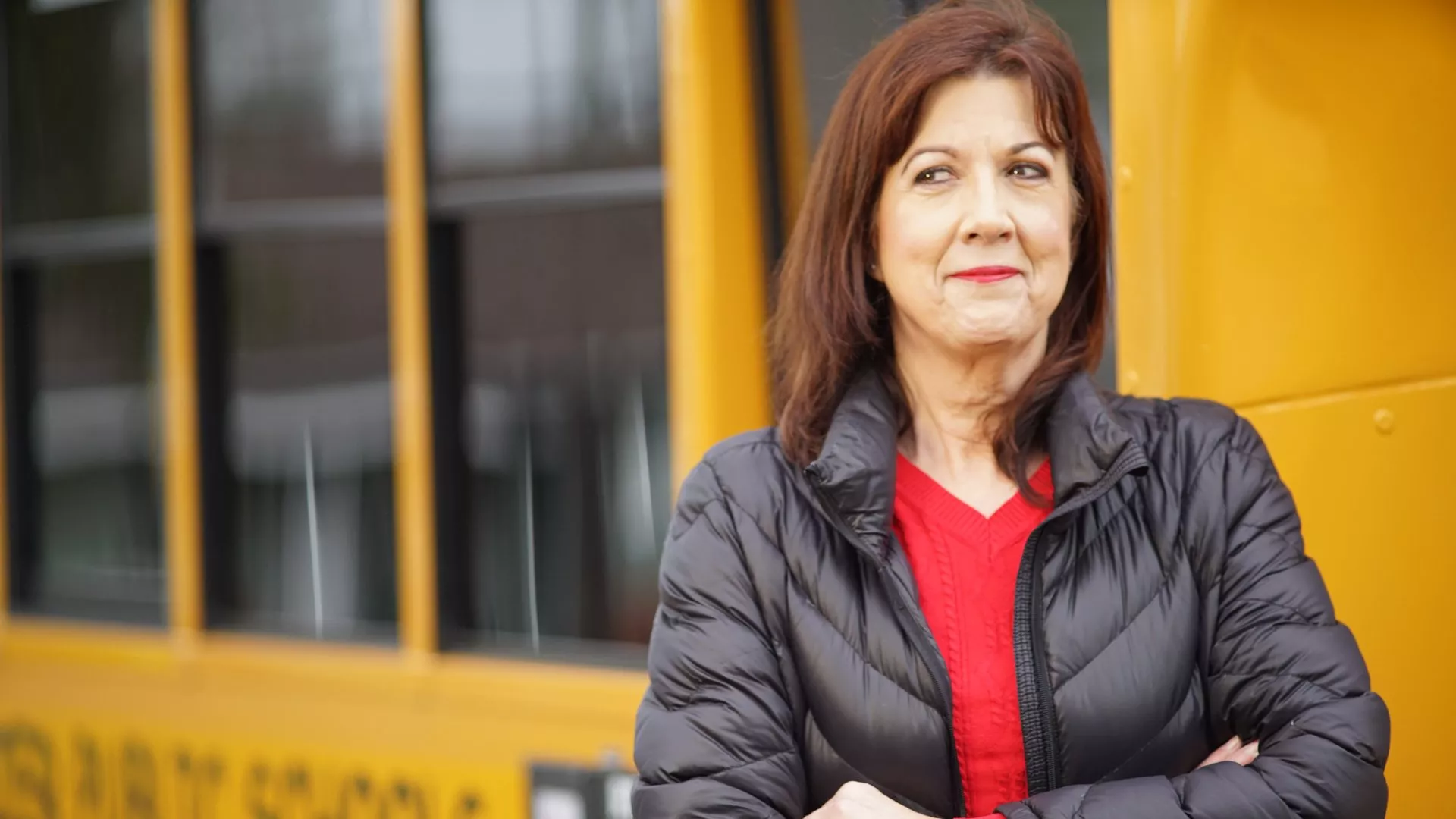 A bus driver poses in front of a bus with a black puffer coat and a red shirt.