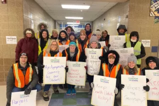 Teachers pose in a school hall with orange vests from working at the crosswalk. They are smiling and holding signs to advocate for honest education standards.